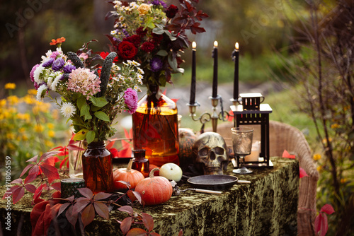 Fall themed holiday table setting arrangement for a seasonal party, glasses, pumpkins, candles, field flowers. Halloween table in the garden with scull, black candles and autumn flowers
