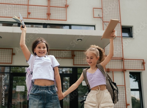 Rear view of multiethnic elementary middle school girls gen Alpha pupils classmates with backpack in casual clothes are hugging embracing each other standing outdoors. Part of a series