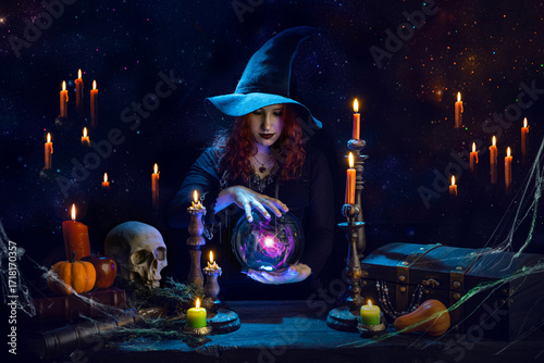Witch casts a spell with magic Crystal ball for predictions fortune-telling. Young woman, magician girl with red hair, dressed in Halloween clothes and conical hat in a room with candles, skull