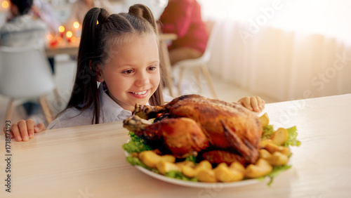 Bilde på lerret Happy little girl looking at roasted Thanksgiving or Christmas turkey, waiting for festive family dinner, her relatives sitting at table on background, copy space