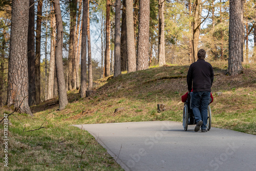 View from back to a woman in wheelchair and a man pushing her in forest park at paved road. Woman with a disability in wheelchair and her personal assistant are walking together