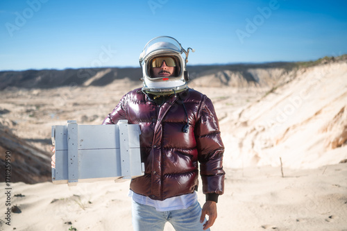 Astronaut wearing helmet holding white wooden box in desert.