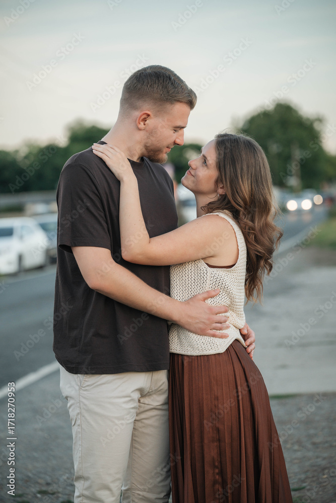Naklejka premium Loving couple embraces on the roadside, gazing into each other's eyes with affection, enjoying a romantic moment outdoors at sunset