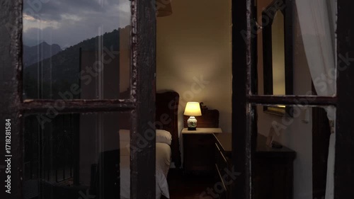 Intimate view of a vintage hotel bedroom through a dark wooden window frame. A warm lamp illuminates the classic antique furniture