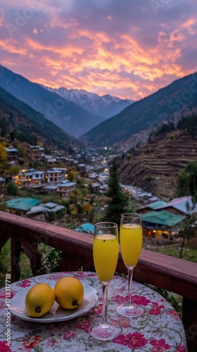 Two glasses of a vibrant yellow drink sit on a table overlooking a mountain village at sunset, with ripe mangoes nearby.