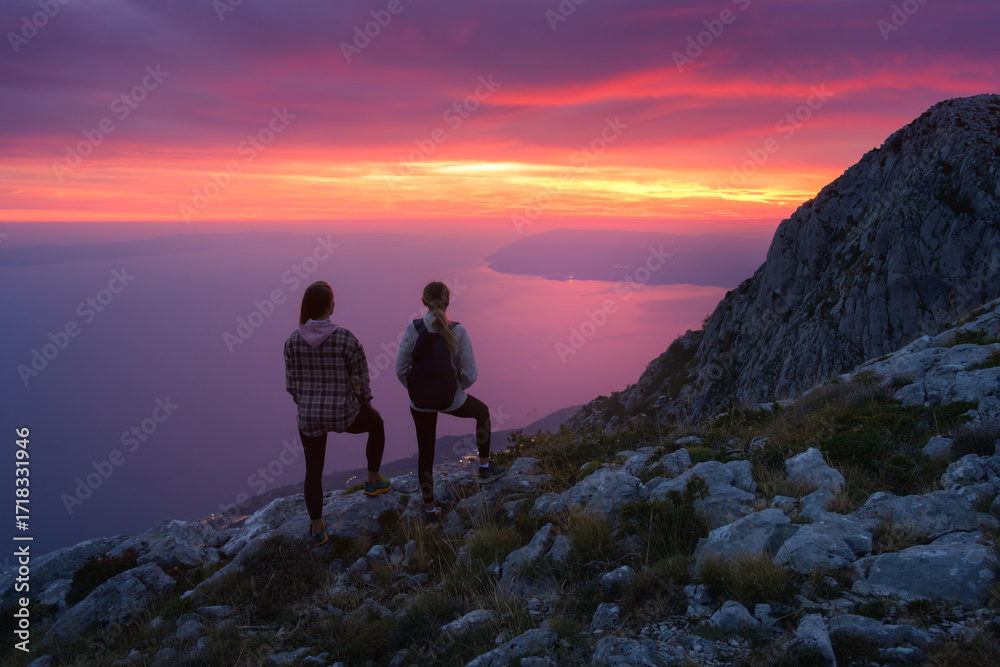 Fototapeta premium Two women hikers with backpacks standing on rocky mountain peak during sunset in Italy, overlooking the sea with dramatic colorful pink sky. Outdoor travel adventure. Sporty girls on mountain at dusk