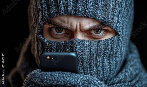 Close-up of a person wearing a balaclava, holding a phone.  Intense, angry expression