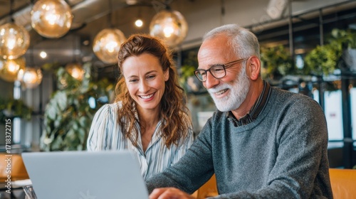 Happy elderly man and young woman using laptop together in modern cafe setting.