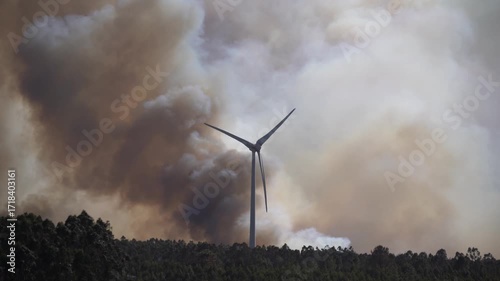 A dramatic wildfire burns near a windmill under a sky filled with thick clouds. The contrast between renewable energy and destructive flames highlights themes of climate change, environmental crisis