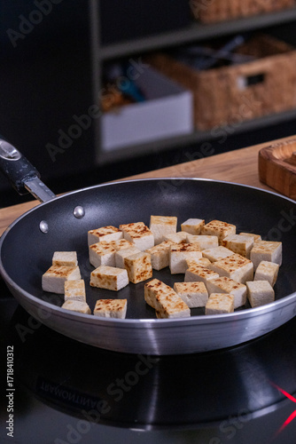 Cubes of golden-brown tofu are sizzling in a non-stick frying pan on a modern stovetop, showcasing the cooking process and vibrant kitchen environment
