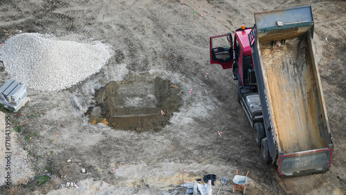 Obraz na plátně Aerial view of construction site with a red dump truck unloading sand and gravel, industrial work in progress, building foundation preparation, heavy machinery and raw materials on ground
