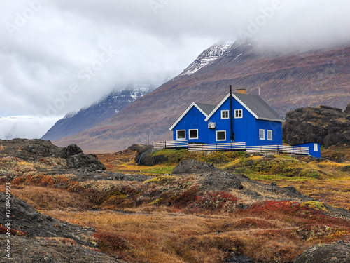 Bright blue house in wild autumn terrain with volcanic rocks and fog-covered mountains in the background.