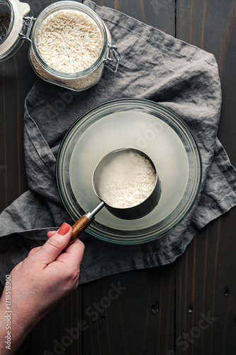 Tablou pe pânză Hand Holding a Measuring Cup of Soaking Sticky Rice: Scoop of glutinous rice hel