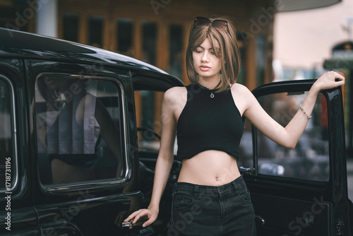 Beautiful young woman in a stylish black outfit stepping into a vintage black car outdoors. The fashionable portrait of a pretty teenager in a retro style.