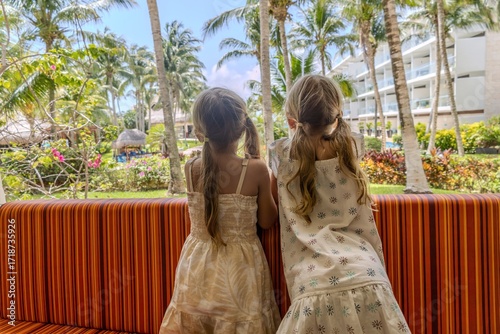 Two girls look out the window at a tropical garden with palm trees. Dressed in summer clothes, they enjoy their vacation at a hotel in a tropical resort. Back view. Family summer vacation