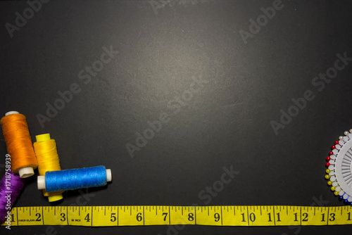 A sewing and tailoring flat lay with colorful thread spools, a measuring tape, and sewing pins on a dark background with ample copy space