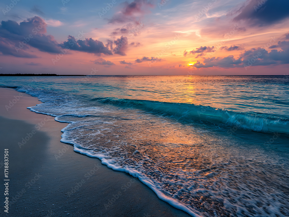 Obraz premium Sunset over ocean waves gently rolling onto sandy beach under colorful sky with clouds creating peaceful atmosphere