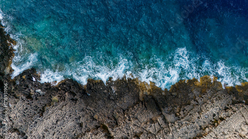 Overhead photo of waves hitting rocky coast with white spray turquoise sea and textured cliffs showing power of ocean against stone surface