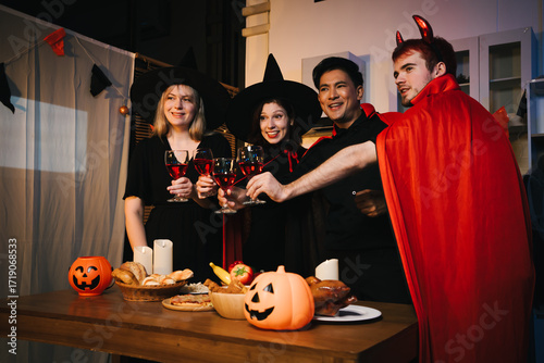 A cheerful group of friends dressed as witches and devils celebrate Halloween together,raising glasses of wine around a festive table with pumpkins,candles, bread,decorations in  cozy indoor setting