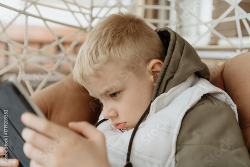 A boy with hearing aids in a jacket plays intently with a tablet on a cozy porch. Captures modern childhood, adaptive technology, and inclusive digital play in a natural setting