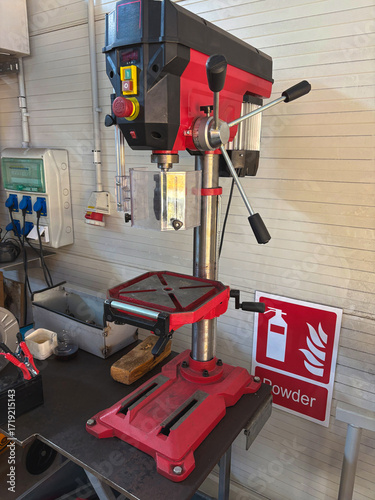 A close-up view of a red and black drill press machine in a workshop, with a sturdy metal base, adjustable table, and safety guards, ready for precise drilling tasks.