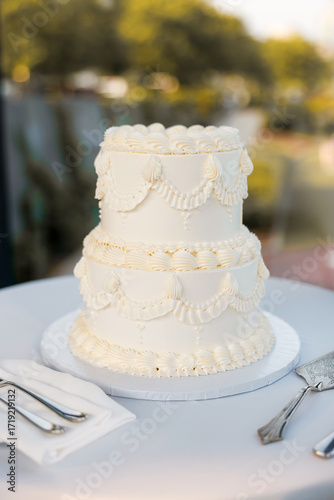 Appetizing cream festive cake at an outdoor wedding banquet. Delicate small white wedding cake decorated with cream patterns. Delicious wedding dessert ready to be cut by the newlyweds