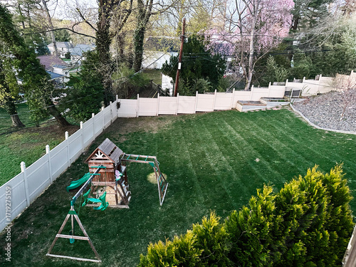 Tablou pe pânză Backyard family playground in USA with white fence, green lawn, and wooden plays