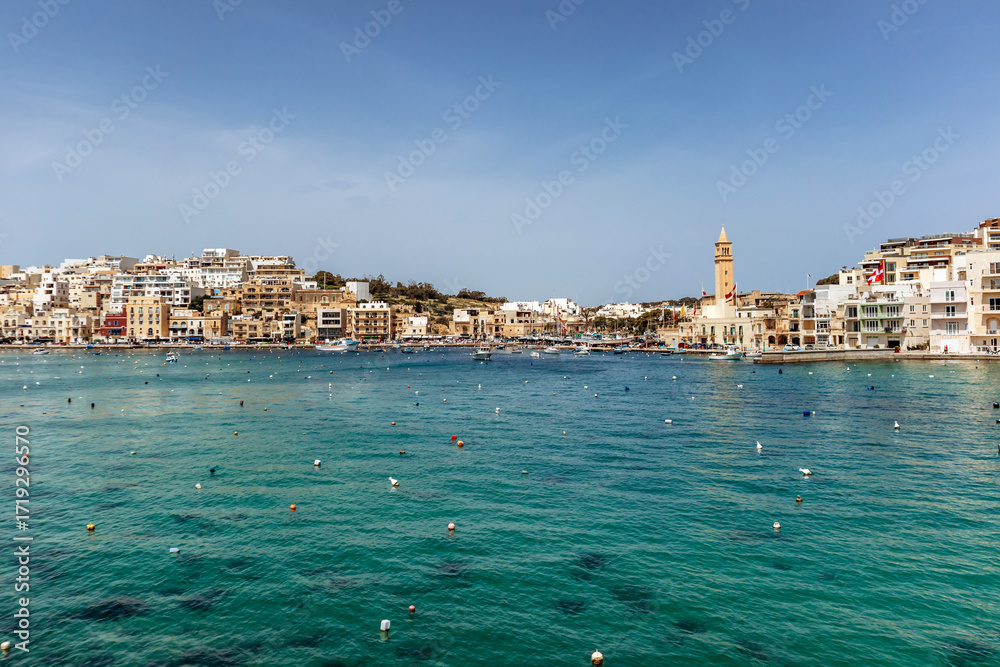 Naklejka premium Panoramic view of the Marsaskala waterfront with colorful buildings, boats, and clear turquoise water on a sunny spring day, Malta, Europe