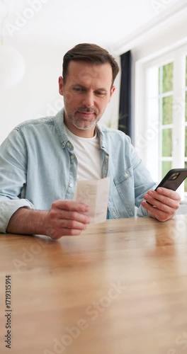 Vertical video of adult man managing home finances using mobile phone holding invoices