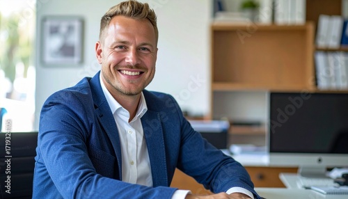 a smiling thirty blonde man with short hair wearing a blue jacket and a white shirt sitting at the desk in the office