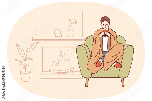 Man feels comfort and harmony sitting by fireplace with blanket on shoulders and cup of hot coffee in hands. Guy enjoys comforts of home and apartment is cozy, allowing him to relax on winter evenings