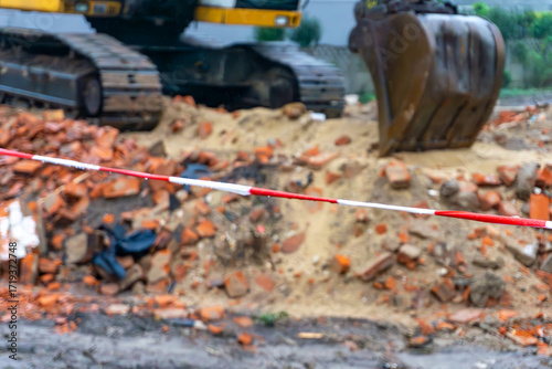 Building demolition site with brick rubble and soil secured by warning tape, crawler excavator with bucket visible in background