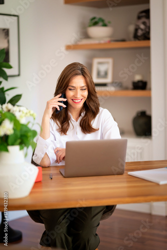 Happy middle-aged woman sitting at home and using smartphone and laptop