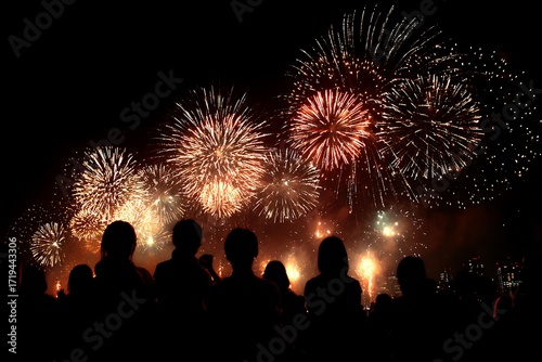 A crowd of silhouetted people watching a magnificent red and gold fireworks show over the city at night