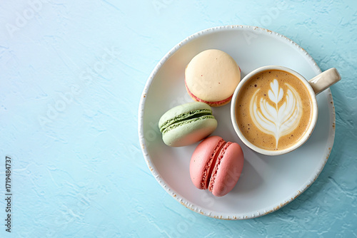 Multicolored French macarons on a plate, next to a cup of cappuccino with a pattern on a light blue vintage texture background. Top view.