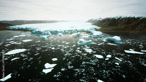 Fotografi Icy giants break apart and tumble into dark waters, surrounded by rugged mountain peaks under a cloudy sky