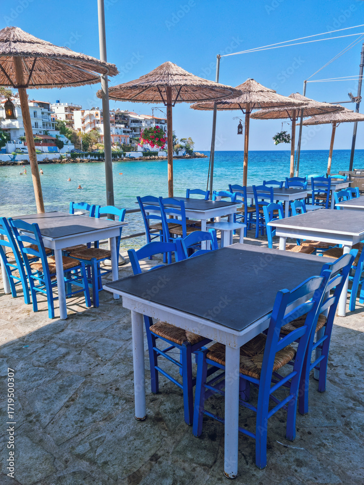 Fototapeta premium Blue Restaurant tables from a traditional Greek taverna overlooking the Mediterrenean sea with turqoise blue waters in the background, Skala Marion, Thassos island, Northern Greece 
