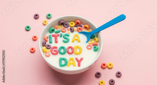 Photo of colorful letter cereals spell out its a good day in a bowl of milk with a blue spoon and scattered cereal pieces on a pink background
