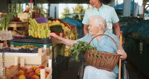 Elderly grandmother in wheelchair choosing fresh tomatoes at farmers market stall, assisted by smiling caregiver in uniform, concept of care, independence, healthy lifestyle and support.