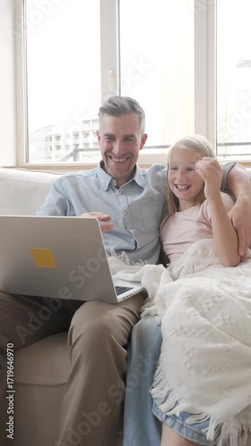 Smiling father and daughter sit on a cozy sofa under a blanket, hugging and making a video call on a laptop. Bright daylight and modern home vibe. Great for family, parenting and connection