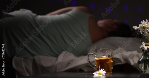 A woman is in her bedroom, resting with dim night lighting. There's a cup of chamomile tea and some fresh Daisies on a table. She is relaxing before bed.