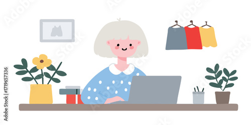 A stylish woman is seated at a desk, engrossed in work on her laptop, with a room for creativity and work. A picture of a shirt, plants in pots and clothes hanging are arranged on the desk. 
