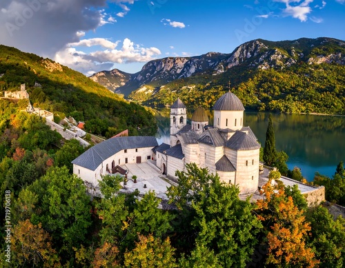 Aerial view of a monastery nestled in a valley, surrounded by autumn foliage and a lake, bathed in golden light