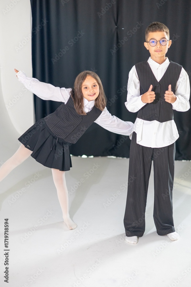Fototapeta premium Two children, a girl and a boy, pose playfully in coordinated formal outfits with playful expressions against a simple backdrop.