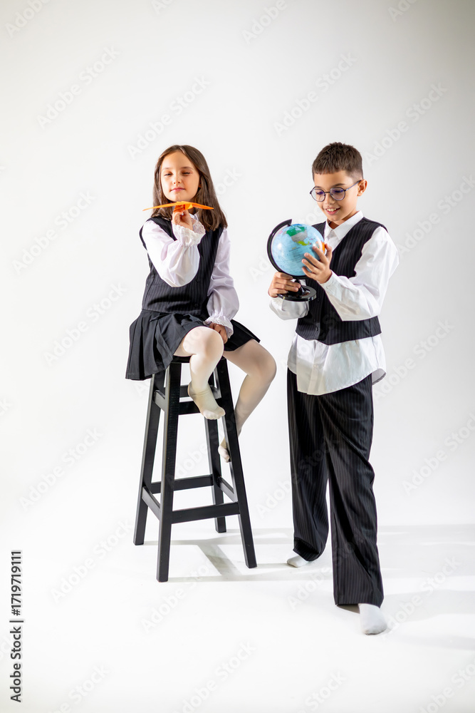 Obraz premium A young Caucasian girl sits on a stool, while a boy stands next to her, exploring a globe. Both wear formal attire, showcasing playfulness and curiosity.
