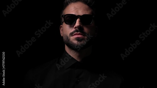 sexy young man with sunglasses in black shirt looking to side, adjusting sunglasses and walking in front of black background