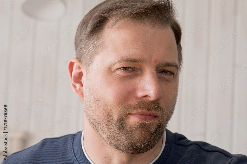 Fototapeta premium Handsome young man in blue jersey indoors. Male with stylish haircut, beard feel happy, calm and serious. Home interior background. Man looking to camera and into distance. Diversity People. Portrait
