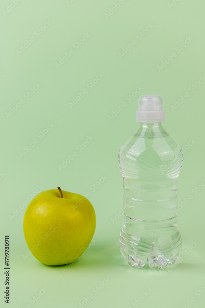 Fototapeta premium Two water bottles, one clear and one green, with a fresh green apple on light green backdrop inspiring wellness