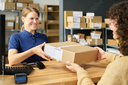 Caucasian young adult woman smiling while handing package to customer across counter in shipping or postal service office, shelves with parcels visible in background, barcode scanner on desk
