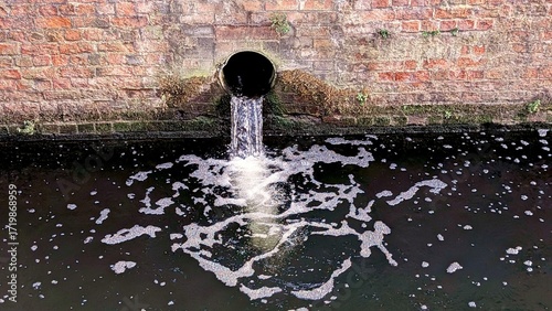 A drainpipe on a brick wall discharging water into a river or canal, symbolizing urban infrastructure and water management